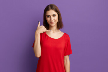 Fototapeta premium Call me. Confidence young pretty brunette girl in red t-shirt doing phone gesture with hand and fingers like talking on the telephone. Asks you to connect with her. Posing over purple background.
