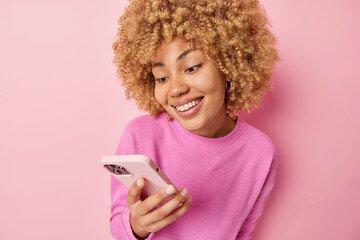 Happy good looking woman with curly hair pleasant smile on face texts on cellular concentrated at screen with glad expression wears casual pullover isolated over pink background. Modern technologies