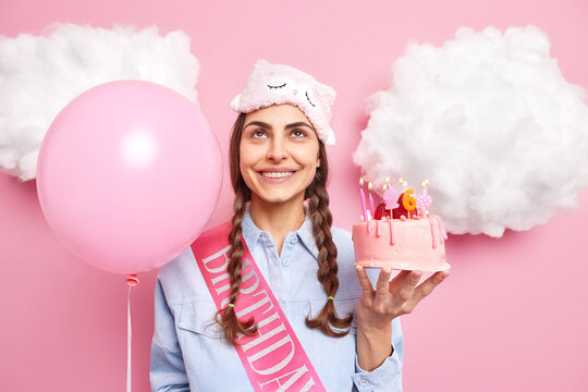 Satisfied Woman With Pigtails Smiles Gently Focused Overhead Holds Appetizing Cake Celebrates Birthday Holds Inflated Balloons Wears Blindfold And Shirt Poses Against Pink Background White Clouds