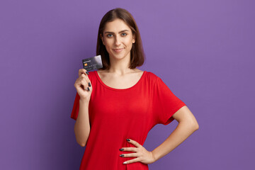 I like cashback. Advertising of your bank. Happy young pretty brunette girl in red t-shirt showing debit or credit card. Posing over purple wall. Using e-banking, e-commerce. Cashless money.
