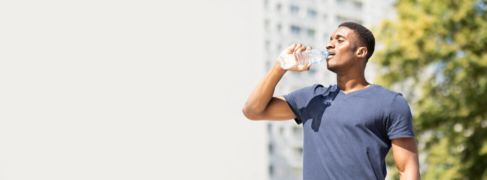 banner sportive young thirsty handsome african american man (guy) drinks water (isotonic) from plastic bottle after cardio workout outside, green tree and building are on background. Healthy lifestyle