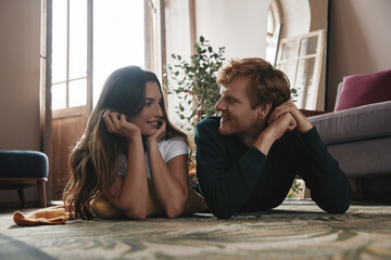 Beautiful young couple looking at each other and smiling while lying on the floor at home together