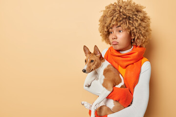 Upset young woman looks sadly aside carries basenji dog returns after walk in bad mood because of bad autumn weather wears white turtleneck and orange scarf around neck isolated over beige background - Powered by Adobe
