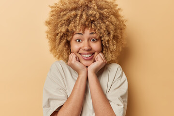 Pretty smiling woman keeps hands under chin shows white teeth dressed in casual t shirt looks directly at camera isolated over beige wall. Positive female model being in good mood hears pleasant news