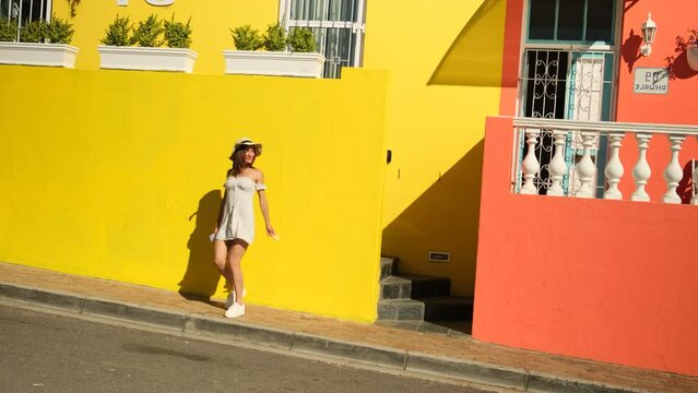 woman in White dress dancing on the street. Joyful african girl in summer dress turning around. stylish girl traveler in a dress holds the flag of south africa and has fun near the colored houses