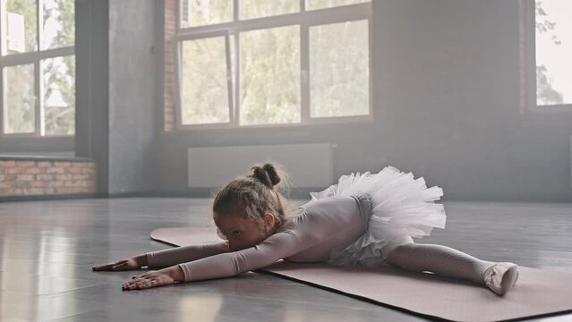Cute little girl does leg-split on beige mat stretching body