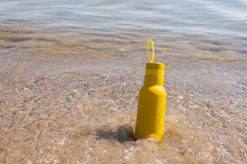 A yellow bottle, thermos standing in the sand on a beach, ocean on sunny summer day. Symbol of...