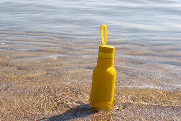 A yellow bottle, thermos standing in the sand on a beach, ocean on sunny summer day. Symbol of summer, holidays and rest