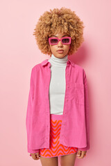 Vertical shot of stylish fashionable woman with curly hair dressed in shirt skirt and sunglasses has serious expression poses against pink wall has self confident face. People clothes style concept