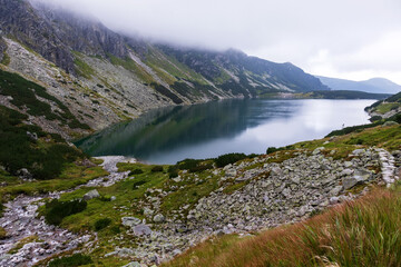 Naklejka na ściany i meble Mountain in the Polish Tatras with a small turquoise lake and rocks in the middle of the mountain with white fog and clouds