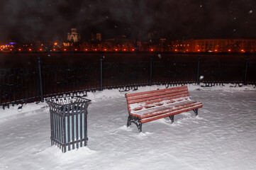A red wooden bench with a black cast-iron urn on the embankment at night. Winter, snowfall - Powered by Adobe