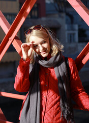 A young beautiful blonde girl stands in a red jacket and covers her face from the sun with her hand. She smiles
