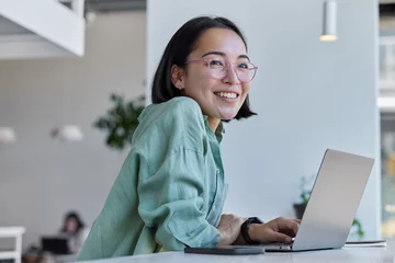 Pretty cheerful Asian woman in eyeglasses and casual clothes browses laptop computer connected to 4g internet updates software uses modern technologies poses in cafeteria looks gladfully away © WHstudio Leushin N