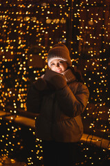 Girl and gift box with Christmas lights. Smiling girl in a hat, scarf looking at the camera at night with blurry lights. Holidays theme