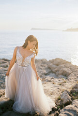 Fototapeta na wymiar Bride in a white puffy dress stands on a rocky seashore holding her hem with her hands