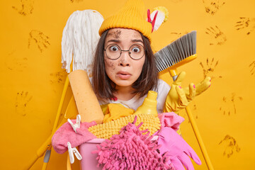 Worried shocked Asian woman stares at camera surrounded by cleaning equipment poses near basket full of laundry and detergents busy doing housework wears hat round spectacles isolated over yellow wall