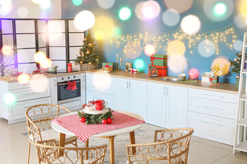 Interior of stylish kitchen with white counters, small Christmas tree, glowing garland and dining table