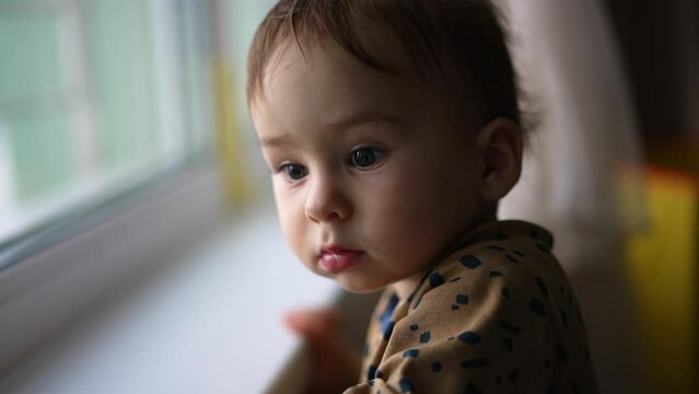 Adorable toddler boy standing at the windowsill. Baby looking curiously at window. Close up. Blurred backdrop.