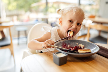 Hungry child girl blond curly with a fork in hand eats with a greedy look into the camera.
