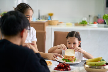 Daughter is eating sandwich on home dinner table with parent