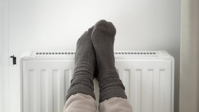 The woman put her frozen feet in woolen warm socks on top of the warm radiator of the home heater. Limiting energy consumption during the cold winter heating period during the global energy crisis.