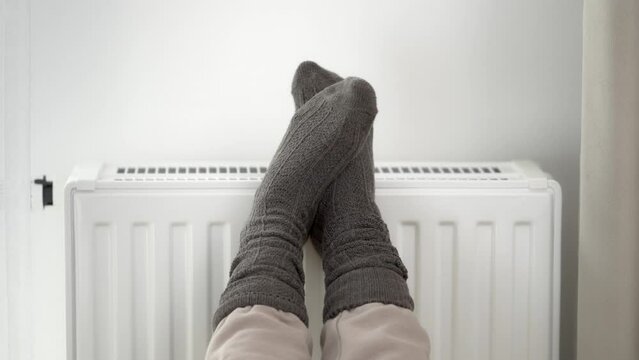 The woman put her frozen feet in woolen warm socks on top of the warm radiator of the home heater. Limiting energy consumption during the cold winter heating period during the global energy crisis.