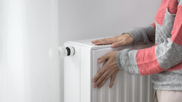 A woman touches the cold radiator of a home heater with her frozen hands and increases the heating power by the maximum value. Limiting energy consumption during the heating season. The energy crisis.