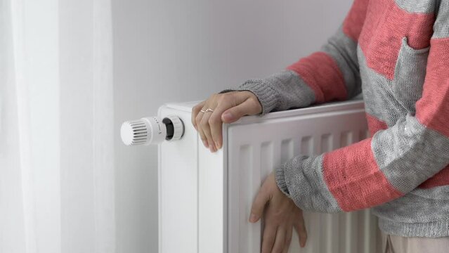 A woman touches the cold radiator of a home heater with her frozen hands and increases the heating power by the maximum value. Limiting energy consumption during the heating season. The energy crisis.