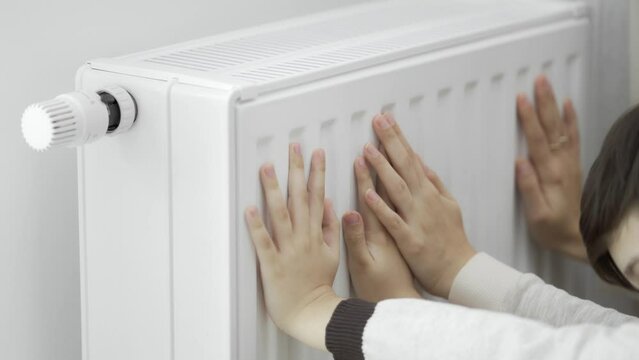 A woman and a child warm their hands by touching the warm radiator of a home heater with their frozen palms in the cold season. Heating of the apartment during the energy crisis.