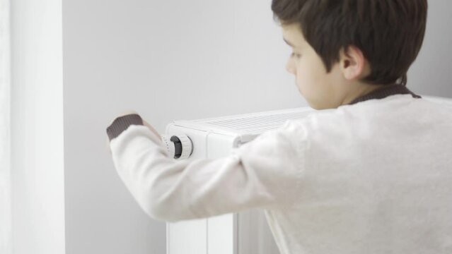 The child touches the cold radiator of the home heater with frozen hands and increases its heating power by the maximum value to warm up. The boy hugs the heater. Heating during the energy crisis.