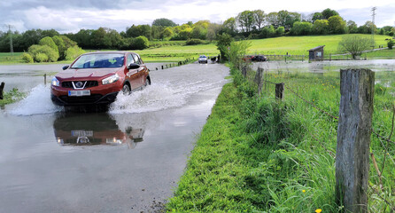 Car driving through deep water puddle on flooded road in Danish countryside due to heavy rain and...