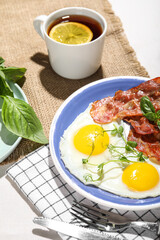 Plate with tasty fried eggs, bacon and cup of tea on table