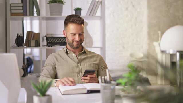 Happy businessman in casual working with phone in home office, Young adult man sitting at desk in study room, writing notes, smiling.