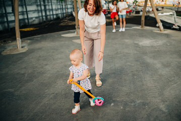 Fototapeta premium Child and mom play together outdoors. Small child on raised arms of mom, play together in city park. Happy family in summer outdoor. Mother and child walk on playground.