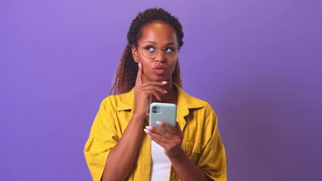 Young positive pensive African American woman with mobile phone holds hand to chin, long time coming up with answer to online test in social network stands in purple studio. Entertainment apps