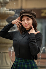 Portrait of a young beautiful dark-haired girl in a cap, a dark shirt and a plaid skirt in the city. Close-up.