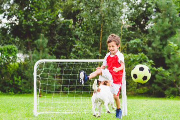 Excited child playing football (soccer) kicks ball while his dog jumping to catch it