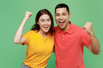 Young happy couple two friends family man woman wear basic t-shirts together do winner gesture celebrate clenching fists say yes isolated on pastel plain light green color background studio portrait.