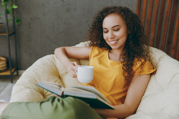 Young happy fun woman of African American ethnicity wear casual clothes drink coffee read novel book sit in armchair stay at home flat rest relax spend free spare time in living room indoor grey wall - obrazy, fototapety, plakaty