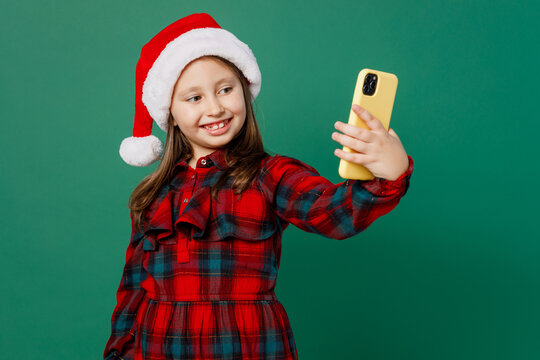 Merry smiling happy little child kid girl 6-7 years old wear red dress Christmas hat posing doing selfie shot on mobile cell phone isolated on plain dark green background. Happy New Year 2023 concept.