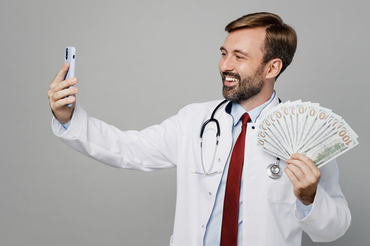 Male doctor man wears white medical gown suit stethoscope work in hospital hold fan of cash money in banknotes do selfie on mobile phone isolated on plain grey background. Healthcare medicine concept.