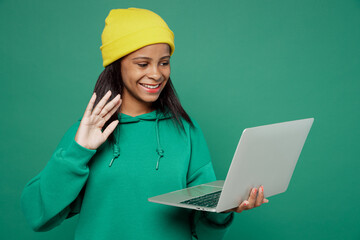 Little IT kid teen girl of African American ethnicity 13-14 years old wear casual hoody hat hold use work on laptop pc computer waving hand get video call talk isolated on plain dark green background.