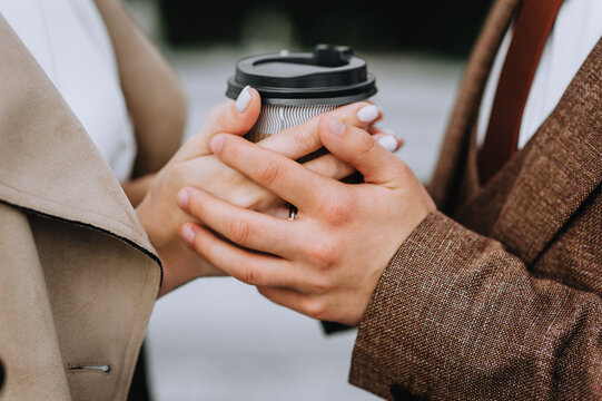 A woman in a coat and a man in a brown suit hold a cup of hot coffee, tea with their hands. Wedding photography of the newlyweds, close-up portrait.