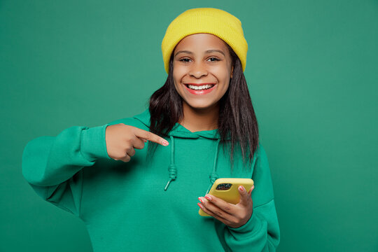 Little kid teen girl of African American ethnicity 13-14 years old wear casual hoody hat hold in hand point finger on use mobile cell phone isolated on plain dark green background Childhood concept. - Powered by Adobe
