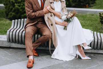 A stylish, fashionable groom in a brown suit gently hugs the bride in a white dress with a bouquet of wild flowers, sitting on a park bench. Wedding photography of the newlyweds, portrait.