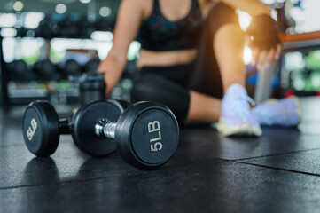 Obraz na płótnie Canvas Dumbbell in the gym with woman blur while sit breaking relax after fitness exercise workout in background. Fitness exercise building muscle of women concept.