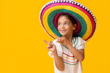 Little Mexican girl in colorful sombrero hat pointing at something on yellow background