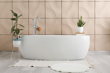 Modern bathtub and houseplants near beige tile wall in bathroom interior