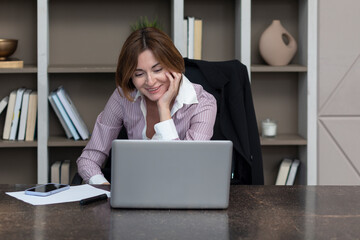 Portrait of successful and attractive business woman working on laptop in her office. Positive female employee  smiling and sitting at the desk in her modern workplace