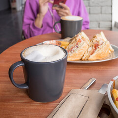 black coffee mug with white foam on the table in a cafe against the background of a woman with a smartphone